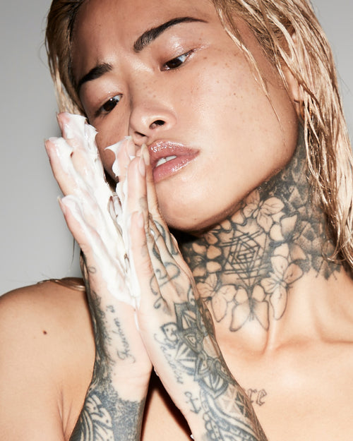 Person with tattoos on neck and arms holding hands near face against a neutral background