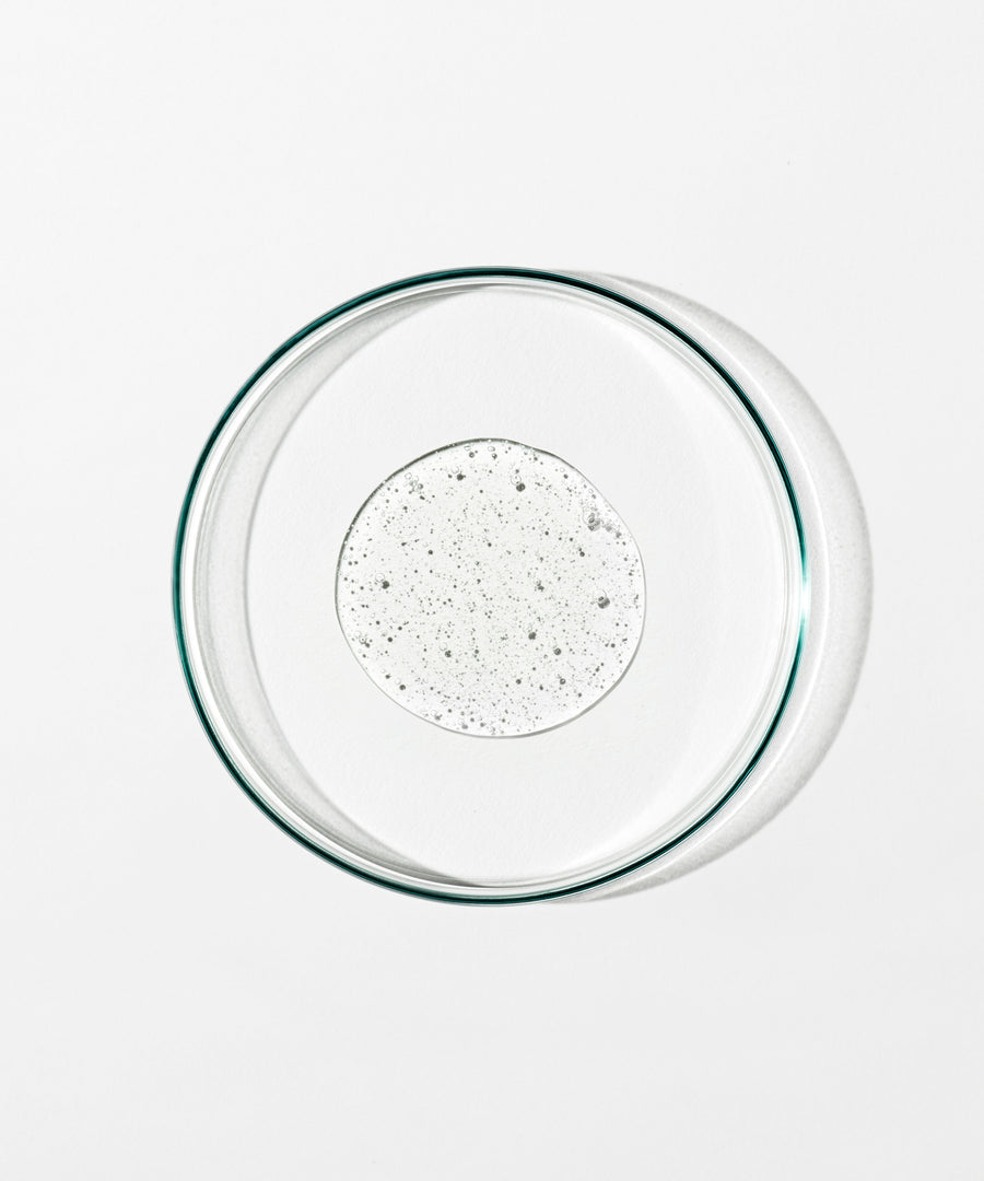 A clear glass petri dish viewed from above, containing a small circular pool of energize body cleanser on a plain white background.