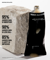 A black squeeze tube of GROWN ALCHEMIS Enzyme Exfoliator stands against a large rough stone on a white studio background, with marketing text on the left describing skin benefits.