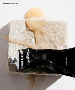 A black squeeze tube labeled GROWN ALCHEMY Enzyme Exfoliator lies across a rough stone slab with a beige exfoliating scrub and a glossy amber gel pool on a white surface.