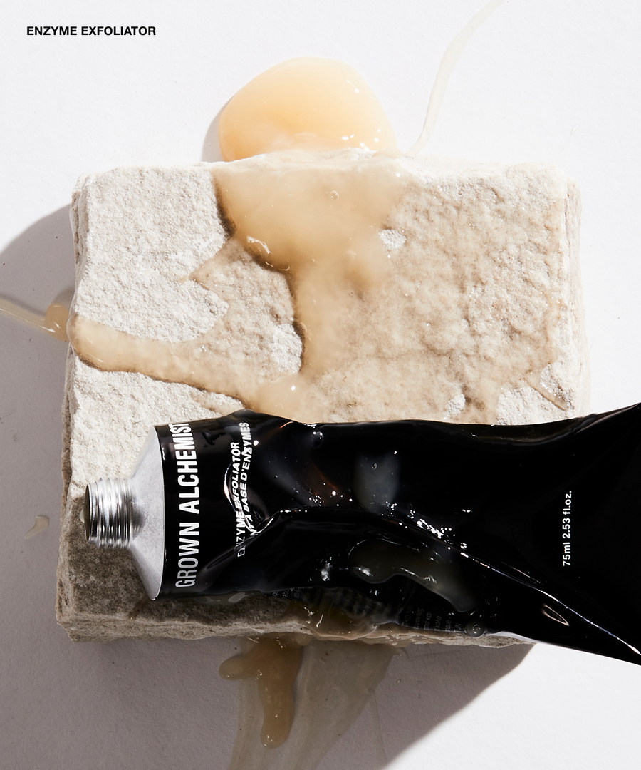 A black squeeze tube labeled GROWN ALCHEMY Enzyme Exfoliator lies across a rough stone slab with a beige exfoliating scrub and a glossy amber gel pool on a white surface.