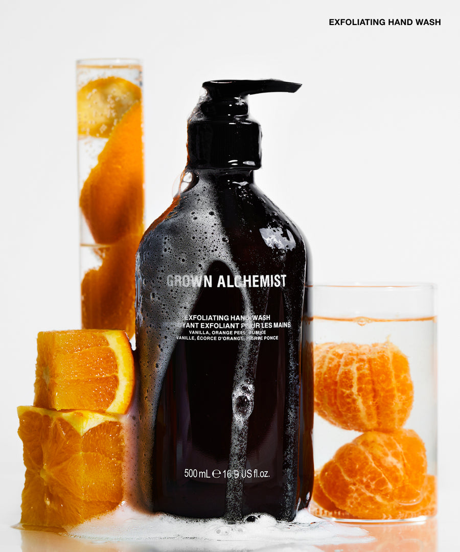 A 500 mL pump bottle of Grown Allchemist Exfoliating Hand Wash stands on a white surface with citrus slices and two glasses of orange-infused water around it. The bottle is dark amber-brown with a black pump and white text.
