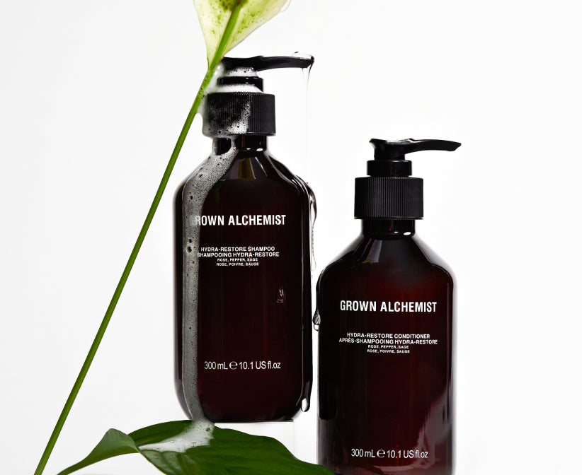 Two bottles of Grown Alchemist shampoo and conditioner with a leaf on a white background