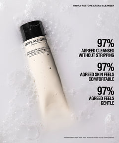 A tube of Brown Alchemy Hydra-Restore Cream Cleanser lies diagonally on white foam. The tube is off-white with a black cap, and bold black text on the right lists user-trial results.