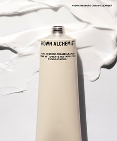A squeeze tube of Grown Alchemist Hydra-Restore Cream Cleanser standing upright against a white background with decorative swirls of the cleanser itself.