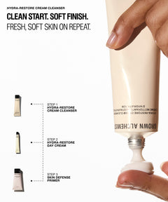 A hand squeezes a cream tube of Brown Allchemists Hydra-Restore Cream Cleanser against a white background with product text and small step-by-step skincare bottle illustrations on the left.