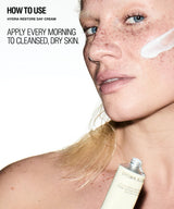 Close-up beauty ad showing a woman with freckles applying or wearing a white cream on her cheek, with text reading 'HOW TO USE HYDRA-RESTORE DAY CREAM' and 'APPLY EVERY MORNING TO CLEANSED, DRY SKIN.' A tube labeled 'BROWN AL.
