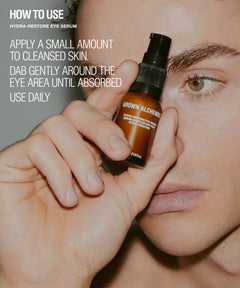 A close-up lifestyle shot of a person holding a small brown bottle of Brown Alchemy Hydra-Restore Eye Serum near their face, with white text on the left explaining how to use the eye serum.