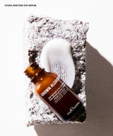 Brown Alchen Hydra-Restore Eye Serum in an amber glass bottle, styled on a textured stone slab next to a white applicator wand, with a clean white background and soft shadow.