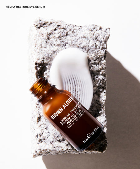 Brown Alchen Hydra-Restore Eye Serum in an amber glass bottle, styled on a textured stone slab next to a white applicator wand, with a clean white background and soft shadow.