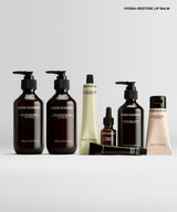 A set of brown and beige cosmetic bottles and tubes from Brow Alchemist, including pump bottles, a dropper bottle, and a squeeze tube, arranged on a light gray background with the text 'HYDRA-RESTORE LIP BALM' at the top right.