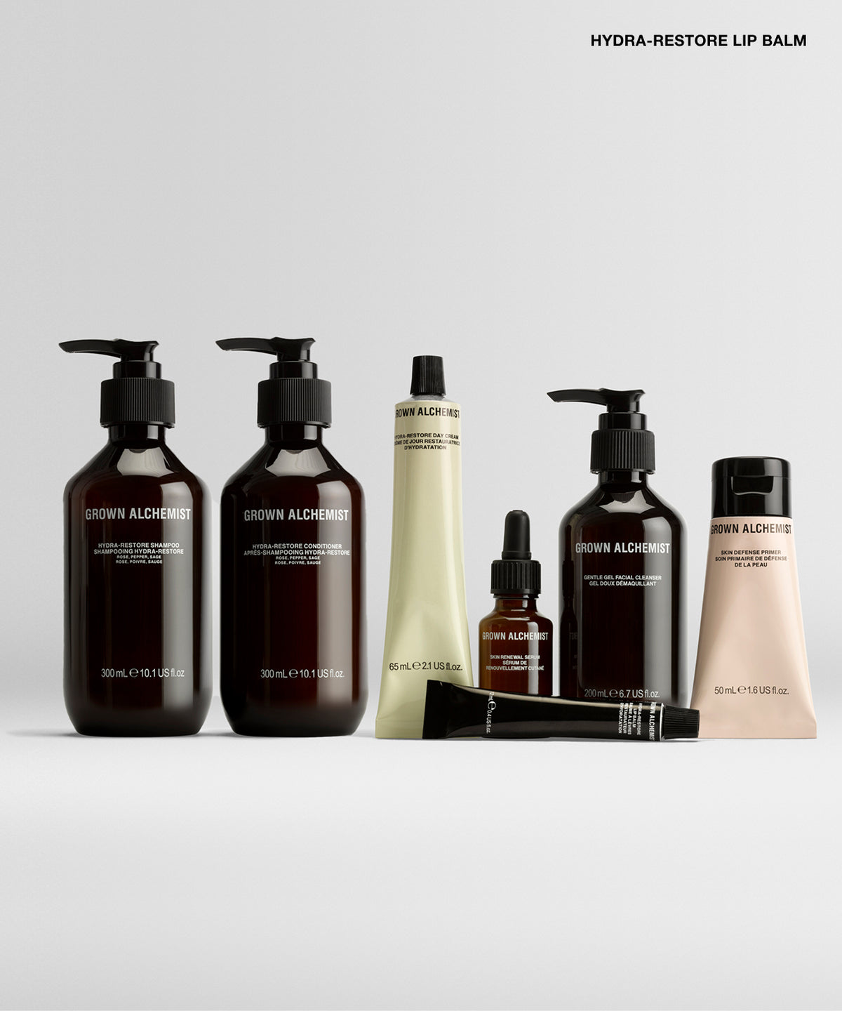 A set of brown and beige cosmetic bottles and tubes from Brow Alchemist, including pump bottles, a dropper bottle, and a squeeze tube, arranged on a light gray background with the text 'HYDRA-RESTORE LIP BALM' at the top right.