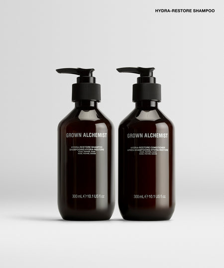 Two amber glass pump bottles of Grown Alchemist Hydra-Restore Shampoo on a light gray background.