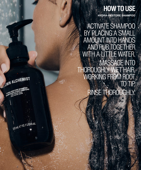 A person with wet dark hair holds a black pump bottle labeled 'Brown Alchemist' while reading a how-to-use overlay for Hydra-Restore shampoo.