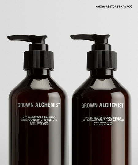 Two dark amber pump bottles of Grown Alchemist haircare products on a light gray background, labeled Hydra-Restore Shampoo and Hydra-Restore Conditioner.