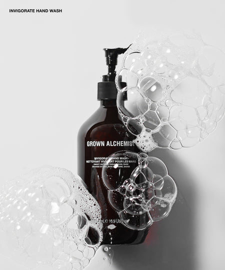 A dark amber pump bottle labeled GROWN ALCHEMIST INVIGORATE HAND WASH, surrounded by large clear soap bubbles on a light gray background.
