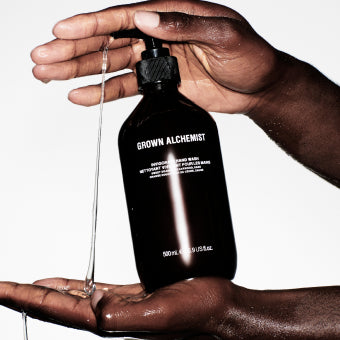 Hand holding a black bottle labeled 'Grown Alchemist' with liquid being poured onto another hand.