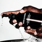 Hand holding a Grown Alchemist bottle with water splashing out, on a white background