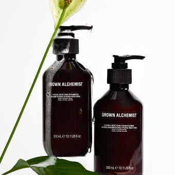 Two bottles of Grown Alchemist shampoo and conditioner with a flower on a white background