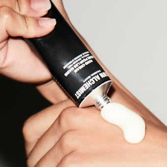 Hand applying Grown Alchemist white cream to a wrist with a black tube against a neutral background