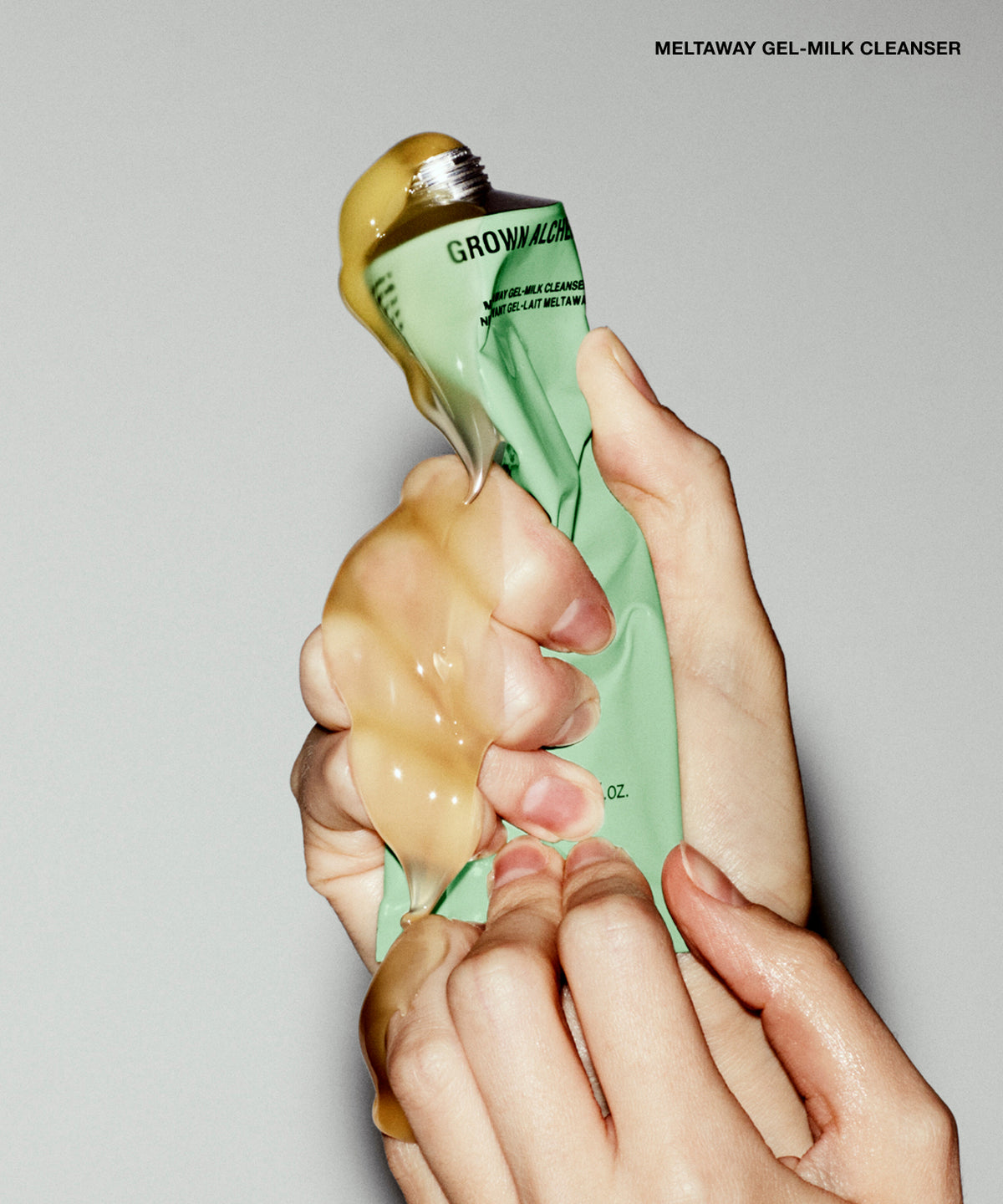 A close-up of hands squeezing a tube of Grown Alchemist gel-milk cleanser, with the thick golden-beige cleanser being expelled against a plain light gray background.