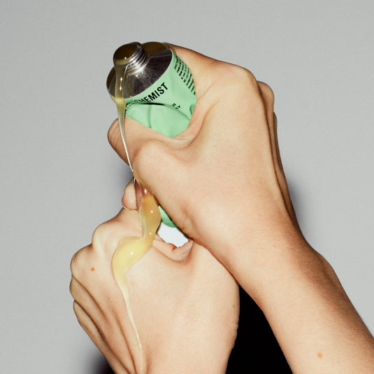 Hand holding a green tube, applying Meltaway Gel-Milk Cleanser to skin against a gray background.