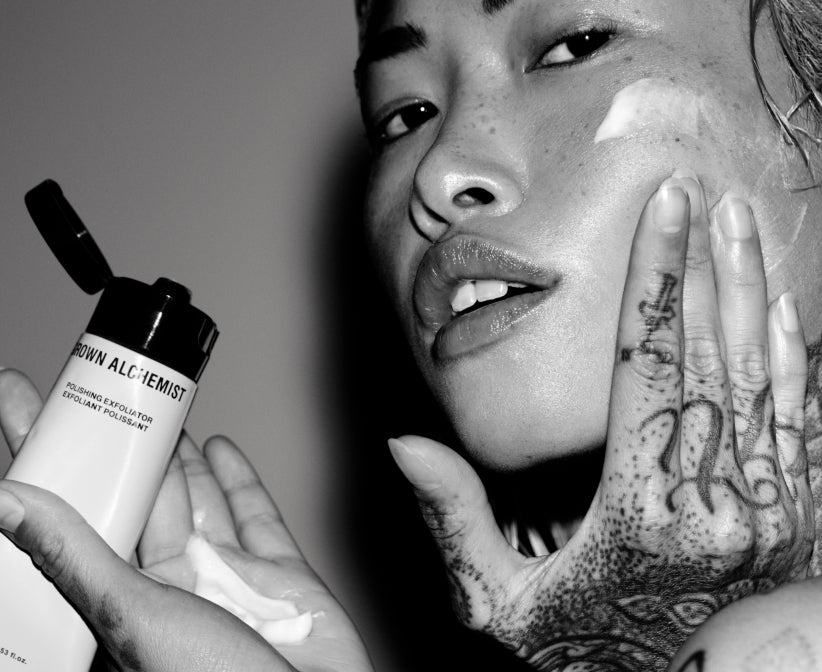 Person applying cream to face with tattooed hand, holding a Grown Alchemist skincare product.