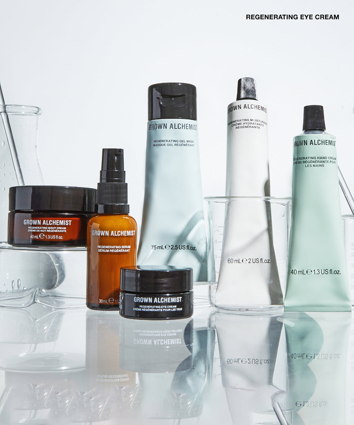 A group of Grown Alchemist skincare products arranged on a reflective white surface with clear glass vessels. Visible products include a large white tube, a pale mint green tube, an amber spray bottle, a dark amber jar, and a black jar, with the text 'REGENERATING EYE CREAM' at the top right.