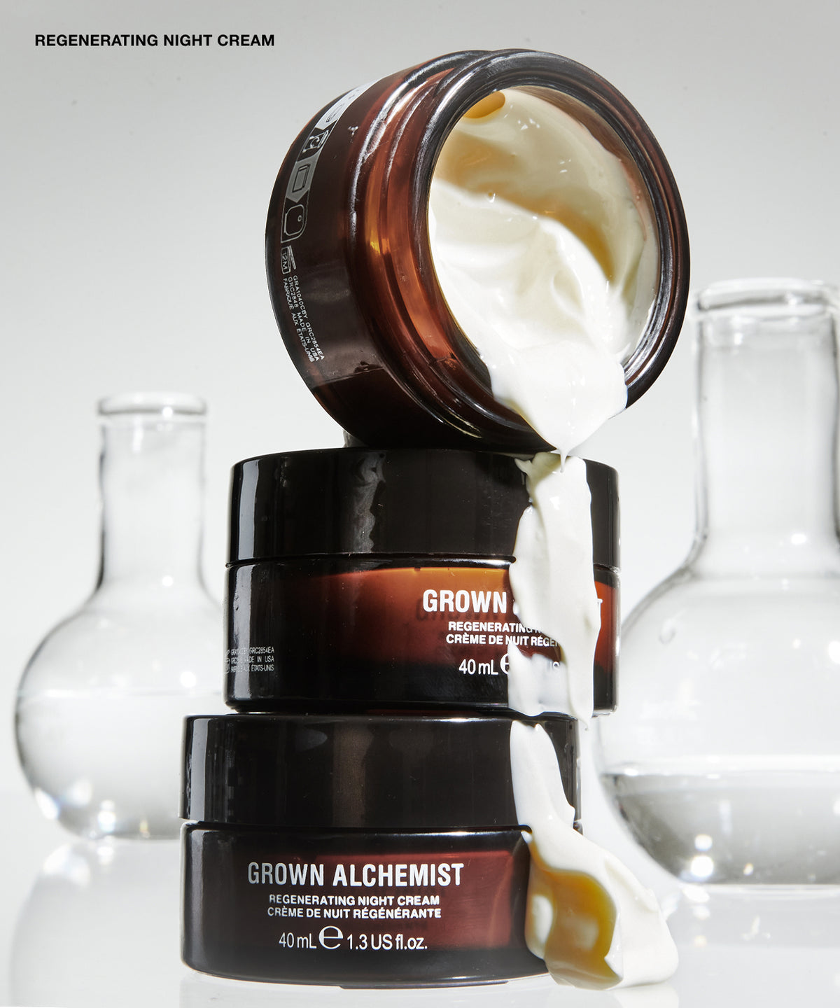 Three cosmetic cream jars stacked and tipped over with thick white cream dripping out, set against a clean light gray background with blurred glass flask shapes. Visible text includes 'REGENERATING NIGHT CREAM'.