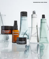 A group of Grown Alchemist skincare products arranged on a reflective white surface with clear glass vessels. Visible products include a large white tube, a pale mint green tube, an amber spray bottle, a dark amber jar, and a black jar, with the text 'REGENERATING EYE CREAM' at the top right.