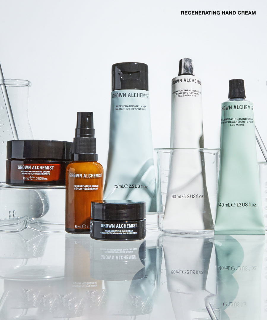 A group of Grown Alchemist skincare products arranged on a reflective white surface with clear glass vessels. Visible products include a large white tube, a pale mint green tube, an amber spray bottle, a dark amber jar, and a black jar, with the text 'REGENERATING EYE CREAM' at the top right.
