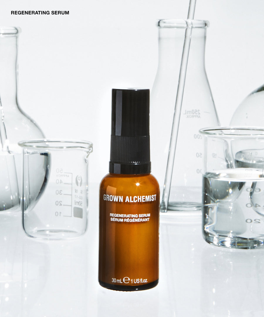 A 30ml amber glass bottle of Grown Alchemist Regenerating Serum with a black spray cap, set against a white background with blurred glass laboratory beakers.