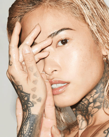 Person with tattoos on hands and face, touching their face against a neutral background