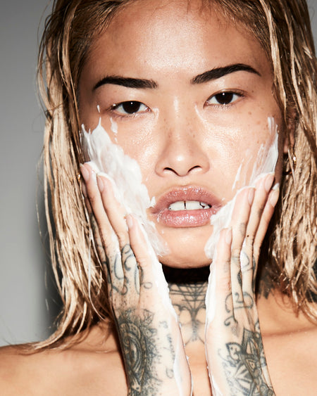 Person with tattoos applying a white cream to their face against a neutral background