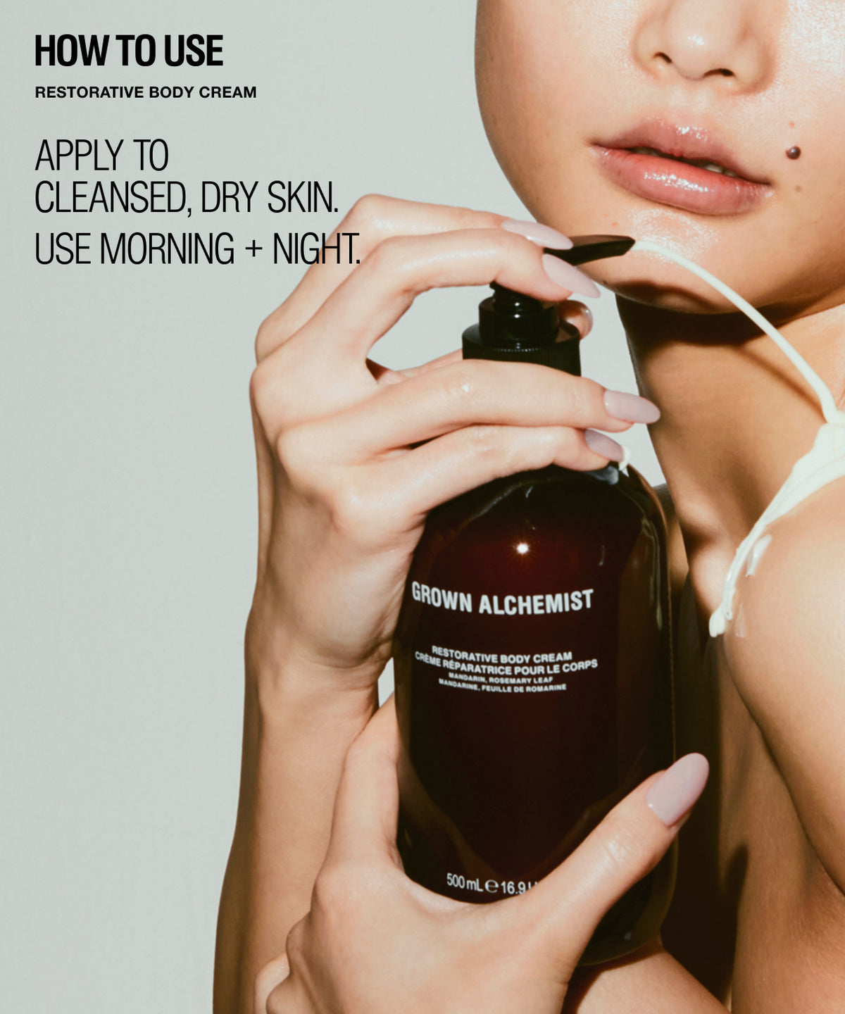 A close-up of a person holding a dark brown pump bottle labeled 'GROWN ALCHEMIST RESTORATIVE BODY CREAM' while white cream is being drawn onto the lips. Text on the left reads 'HOW TO USE RESTORATIVE BODY CREAM' and 'APPLY TO CLEANSED, DRY SKIN.