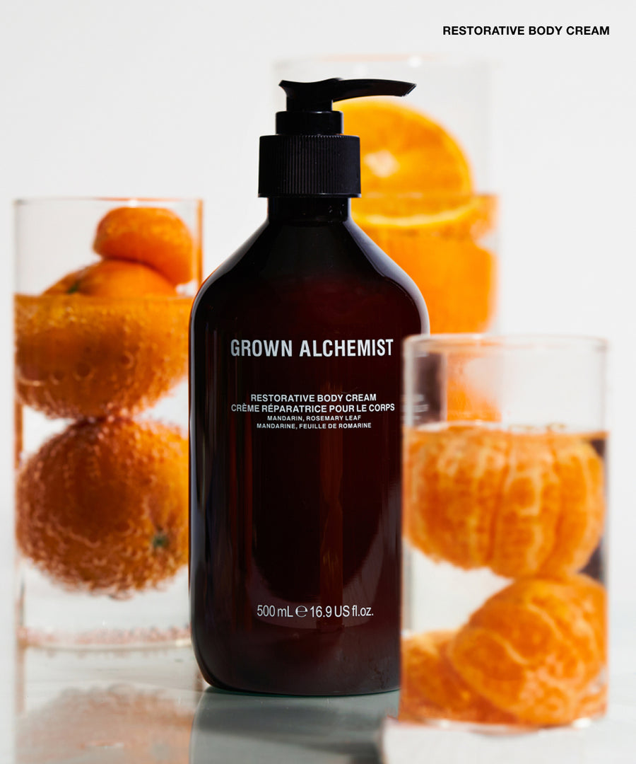 A pump bottle of Grown Alchemist Restorative Body Cream stands centered on a reflective white surface, with orange slices and whole oranges arranged behind it in two clear glasses. The bottle is dark amber-brown with a black pump and white text.
