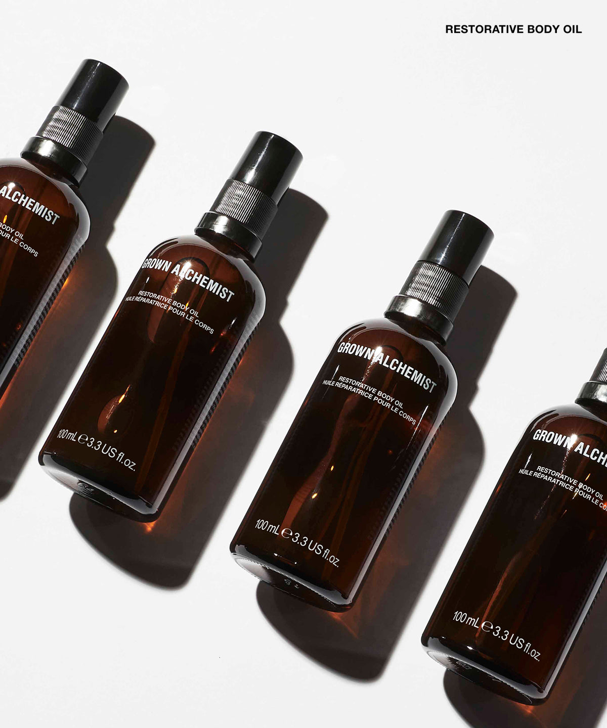 Four amber glass spray bottles of GROWN ALCHEMIST Restorative Body Oil arranged diagonally on a white background with strong shadows.