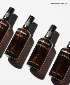 Four amber glass spray bottles of GROWN ALCHEMIST Restorative Body Oil arranged diagonally on a white background with strong shadows.