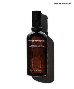 A 100ml amber glass bottle of Grown Alchemist Restorative Body Oil with a black spray pump, cast in a dramatic shadow on a white background.