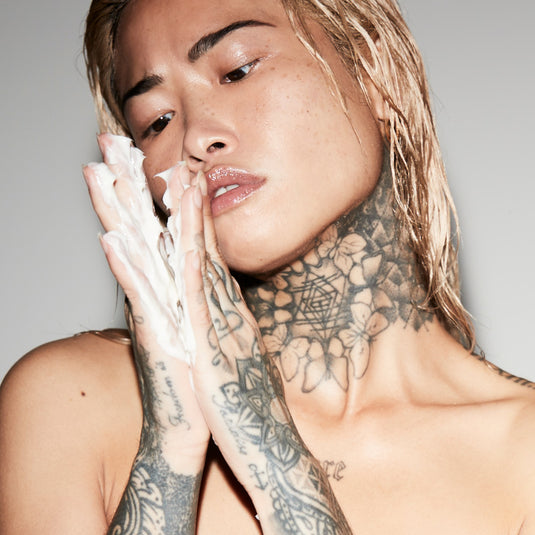 Person with tattoos on neck and arms holding hands near face against a neutral background