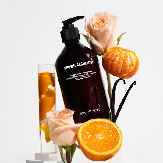 Grown Alchemist hand cream bottle with oranges, roses, and vanilla bean on a light background