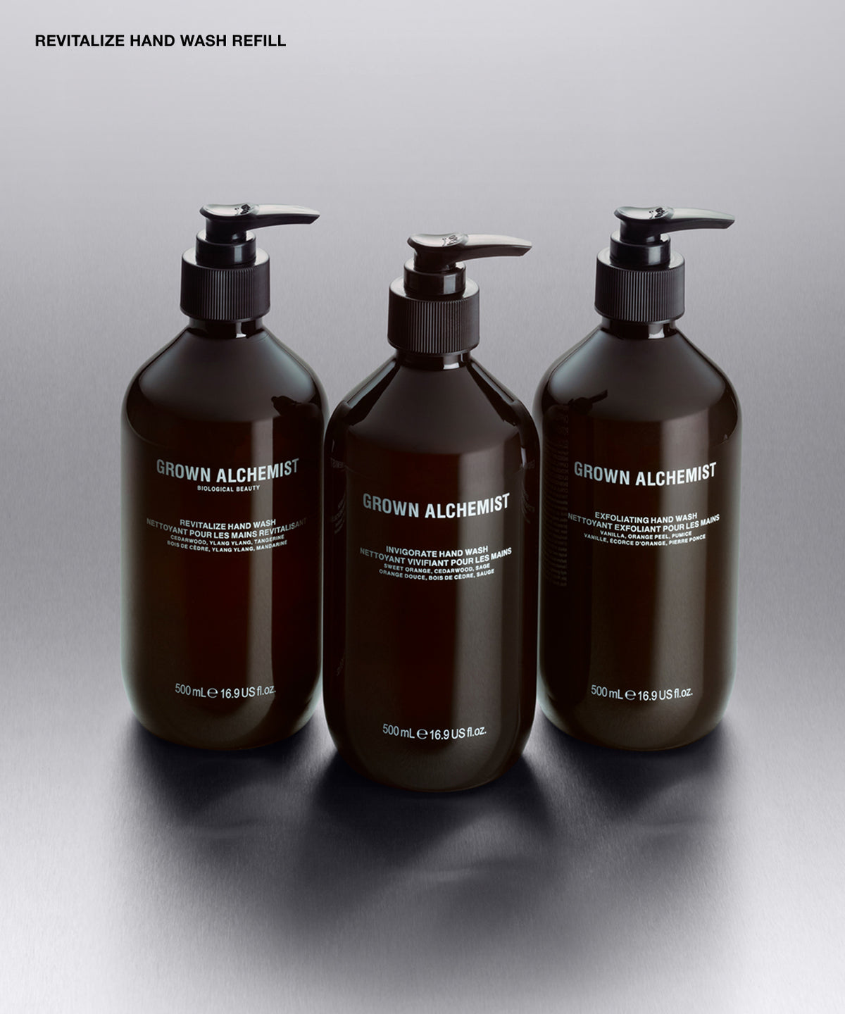 Three amber glass pump bottles of Grown Alchemist hand washes arranged on a reflective gray surface against a dark gray background. The top left corner includes the text 'REVITALIZE HAND WASH'.