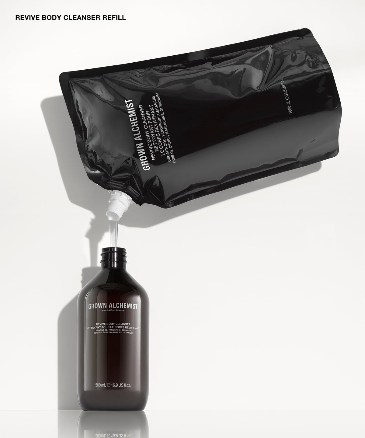 Revive Body Cleanser Refill shown with a glossy black resealable pouch tilted above a dark amber bottle, with white liquid being dispensed from the pouch into the bottle on a clean white background.