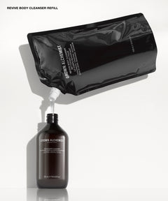 Revive Body Cleanser Refill shown with a glossy black resealable pouch tilted above a dark amber bottle, with white liquid being dispensed from the pouch into the bottle on a clean white background.