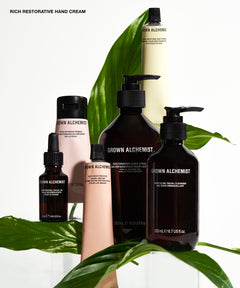 A group of Grown Alchemist hand and body care products arranged with large green leaves on a white background. Visible products include a large dark amber pump bottle labelled Rich Restorative Hand Cream, a smaller dark amber dropper bottle, a pale pink pump bottle, a pale pink tube, and a tall cream-colored bottle.