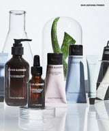 A group of Grown Alchemist skincare products arranged on a reflective white surface, including a large dark brown pump bottle, a small amber dropper bottle, a pale pink tube, a blue-gray squeeze tube, and other containers, with a glass dome and aloe leaves in the background.