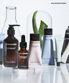 A group of Grown Alchemist skincare products arranged on a reflective white surface, including a large dark brown pump bottle, a small amber dropper bottle, a pale pink tube, a blue-gray squeeze tube, and other containers, with a glass dome and aloe leaves in the background.