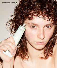 A close-up lifestyle shot of a woman with curly brown hair holding a tube of Skin Renewal Day Cream by La Roche-Posay. The text 'SKIN RENEWAL DAY CREAM' is visible in the top left corner.