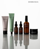 A six-piece skincare set from Serenity Wellness arranged on a reflective white surface against a white background. The set includes a light pink tube, a sage green tube, two amber dropper bottles, an amber spray bottle, and a black jar, with the text 'SKIN RENEWAL FACIAL OIL' in the top right corner.