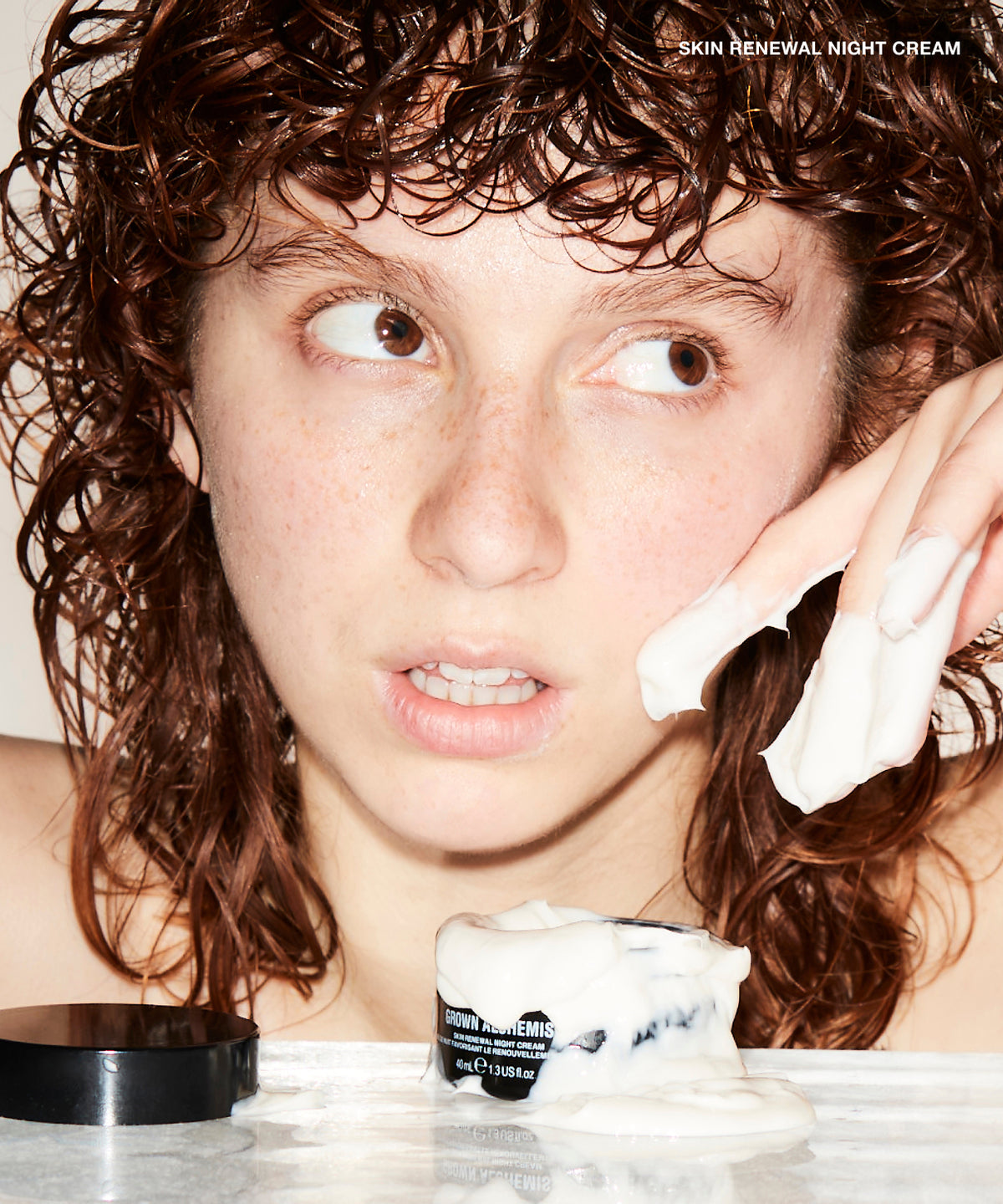 A close-up lifestyle shot of a woman with curly brown hair applying or touching up white cream on her cheek, with an open jar of 'Skin Renewal Night Cream' and its black lid in the foreground.